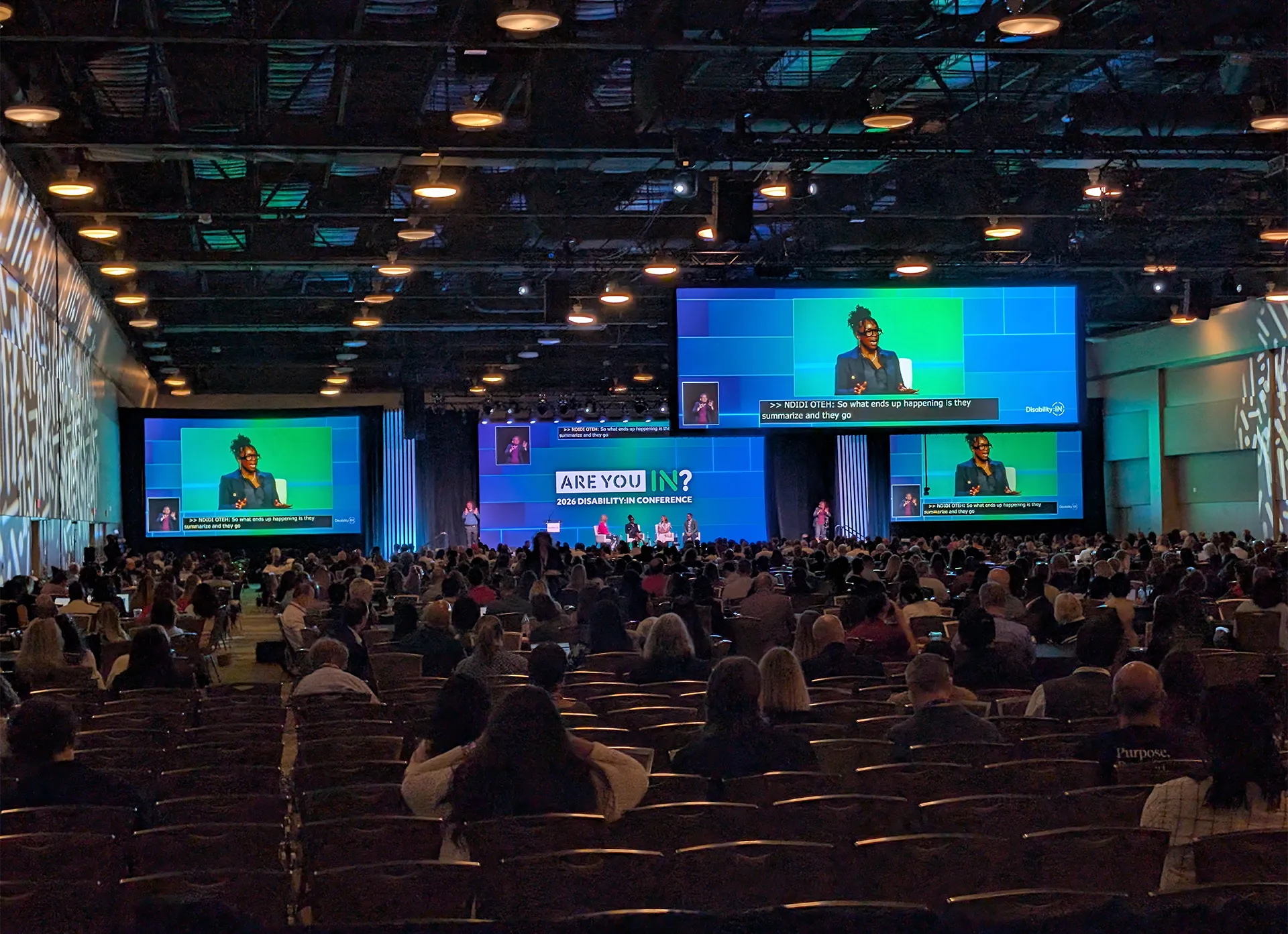 Disability:IN Global Conference presentation with a speaker on stage, accessible captions and sign language interpretation displayed on large screens.